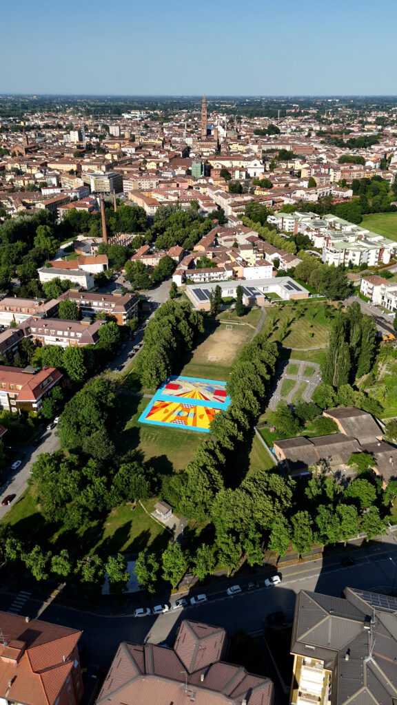 IMMAGINE: Veduta panoramica dall’alto della città. Al centro di un grande parco alberato spiccano i due campi da basket riqualificati, riconoscibili per le superfici dai colori accesi.