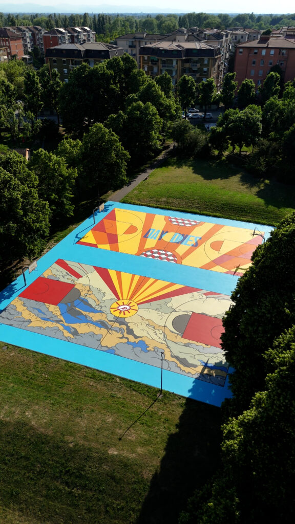 IMMAGINE: Veduta aerea di due campi da basket riqualificati all’interno di un parco urbano. Le superfici combinano motivi geometrici arancioni e gialli, onde, nuvole e raggi solari, incorniciati da un bordo azzurro.