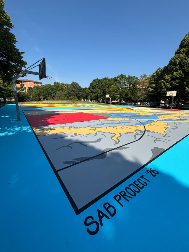 IMMAGINE: Vista dal bordo del campo da basket completato, decorato con onde e nuvole stilizzate nei toni del grigio, beige e blu. La cornice è azzurra e riporta la scritta nera “SAB Project ’26”.