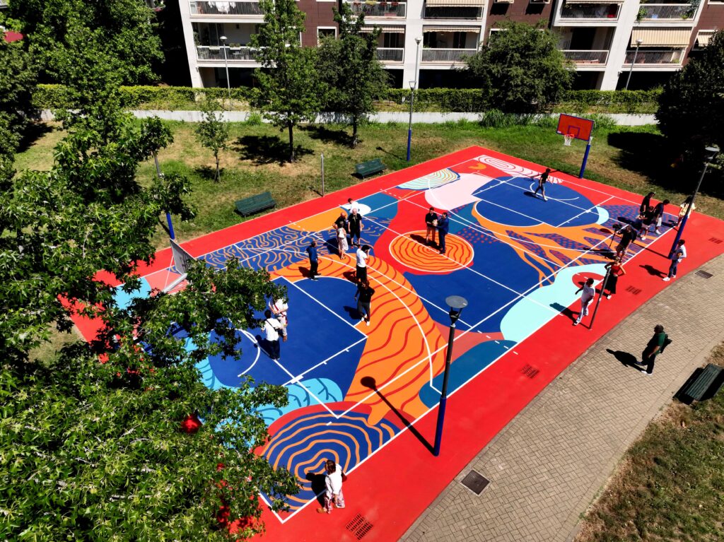 IMMAGINE: Veduta aerea del campo da basket riqualificato durante un momento di apertura al pubblico. La superficie è decorata con grandi forme organiche nei toni del rosso, blu, arancione e azzurro, mentre diverse persone attraversano il playground.