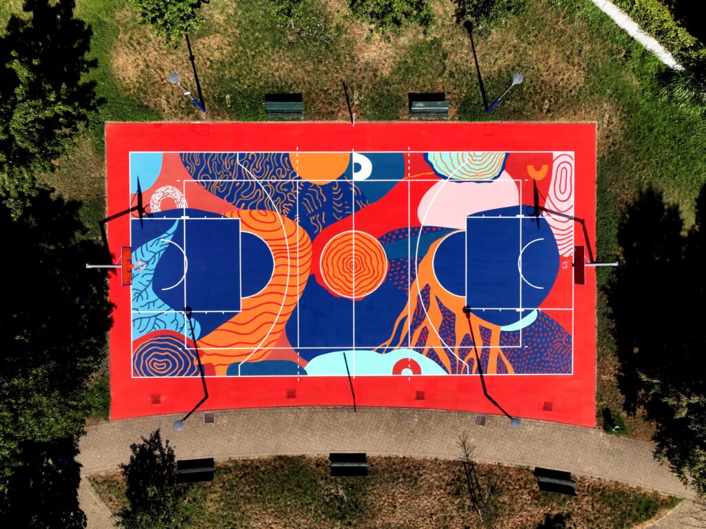 IMMAGINE: Vista zenitale del campo da basket immerso nel verde. La composizione artistica combina forme astratte e motivi naturali in rosso, blu, arancione, rosa e azzurro, attraversati dalle linee bianche di gioco.