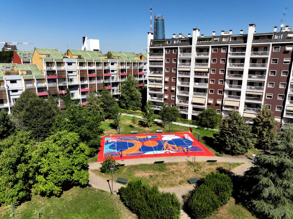 IMMAGINE: Veduta aerea obliqua del campo riqualificato all’interno del parco, circondato da alberi e palazzi residenziali. Il playground presenta una grafica astratta dai colori accesi, incorniciata da una superficie rossa.