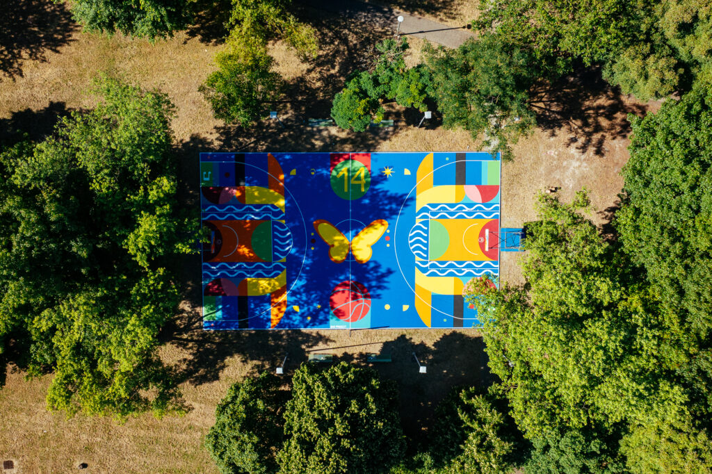 IMMAGINE: Vista dall’alto del campo da basket immerso nel verde. Il playground presenta una composizione simmetrica su fondo blu, con forme geometriche, onde, una farfalla, un pallone e il numero 14.