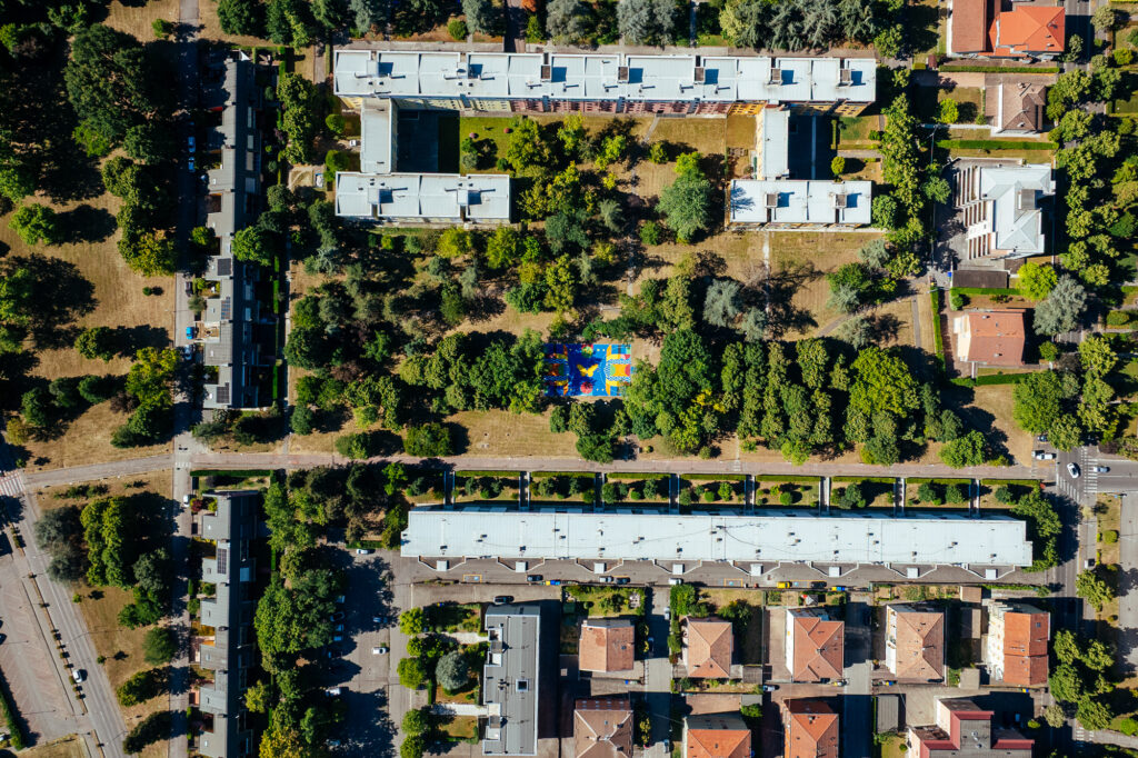 IMMAGINE: Veduta aerea del quartiere e del grande parco urbano. Al centro, tra alberi e edifici residenziali, spicca il campo da basket riqualificato dai colori vivaci.