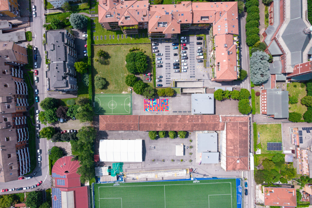 Veduta aerea del quartiere di Colognola: al centro del complesso sportivo e scolastico spicca il campo colorato con la scritta “NOT JUST PHONE”, circondato da edifici, parcheggi e aree verdi.