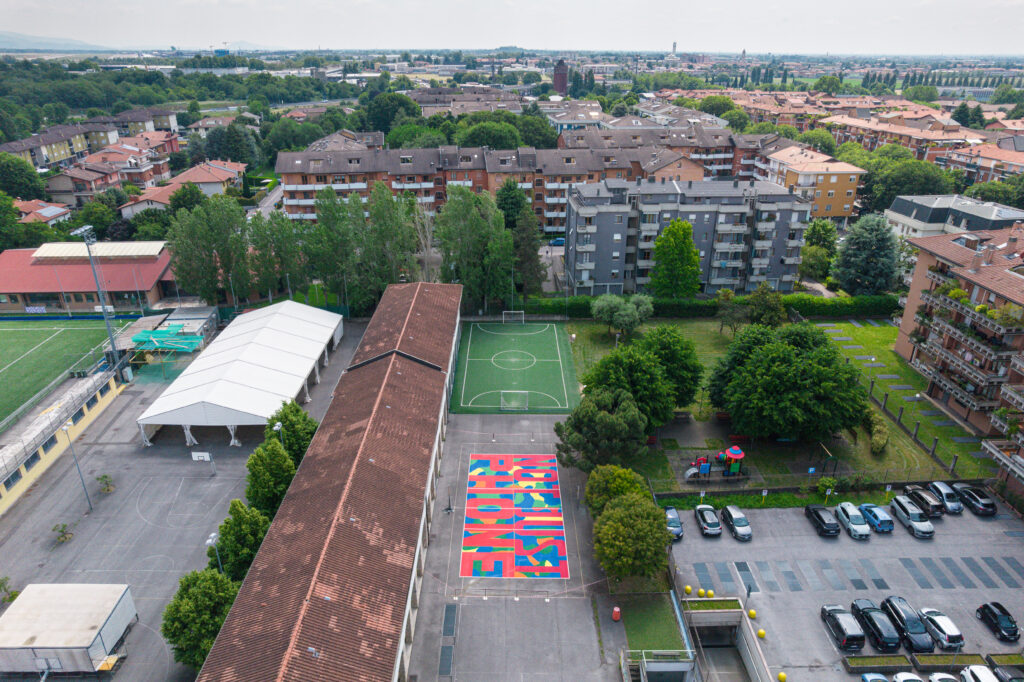 Veduta panoramica dall’alto del quartiere e degli impianti sportivi. Al centro, accanto a un campo da calcio e a un’area giochi, emerge il campo riqualificato dai colori accesi.