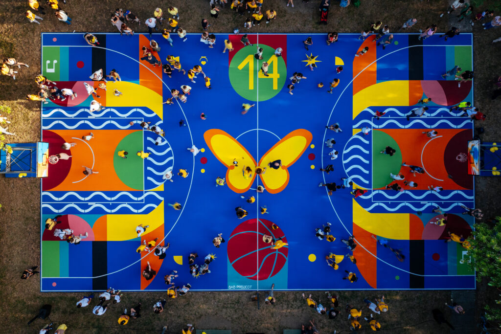 IMMAGINE: Vista zenitale del campo da basket durante l’inaugurazione, affollato da persone. La superficie blu è decorata con forme geometriche multicolori, onde, una farfalla gialla, un pallone e il numero 14.