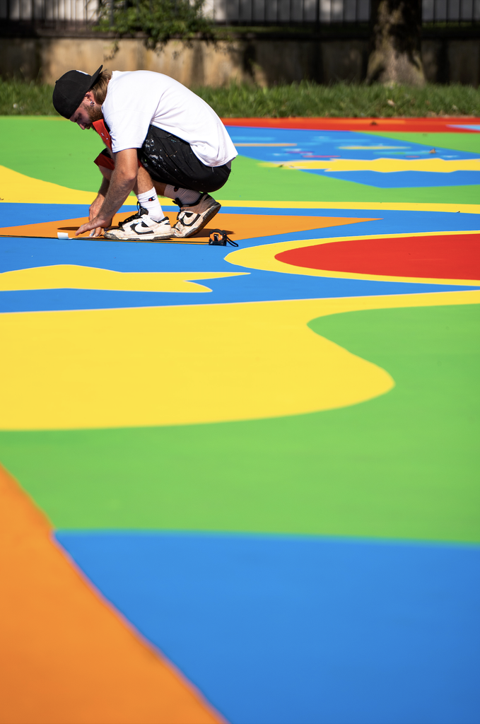 Un artista al lavoro sul playground SAB di Cisano Bergamasco, accovacciato mentre rifinisce con il nastro adesivo una delle forme geometriche colorate dipinte a terra.