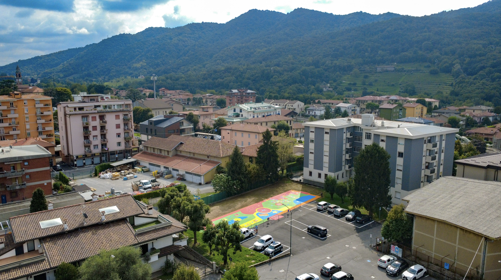 Veduta aerea del nuovo playground SAB di Cisano Bergamasco inserito nel tessuto urbano, accanto a edifici residenziali e a un parcheggio, con le montagne sullo sfondo.