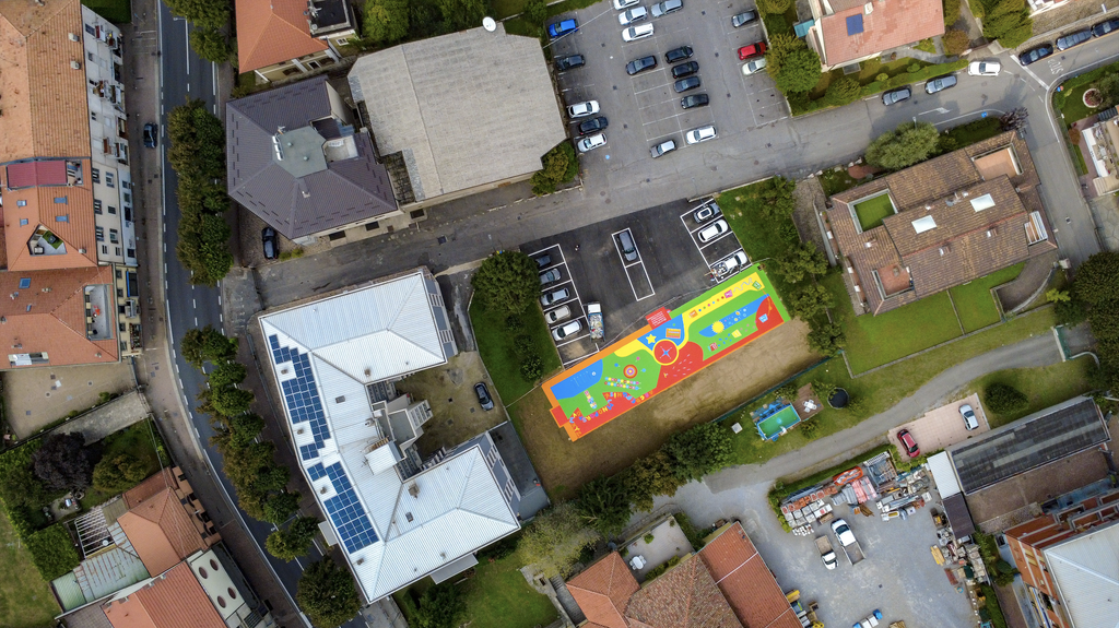 Vista zenitale del playground SAB di Cisano Bergamasco: una lunga superficie rettangolare dai colori vivaci, con percorsi e giochi tradizionali dipinti a terra, circondata da edifici, verde e parcheggi.