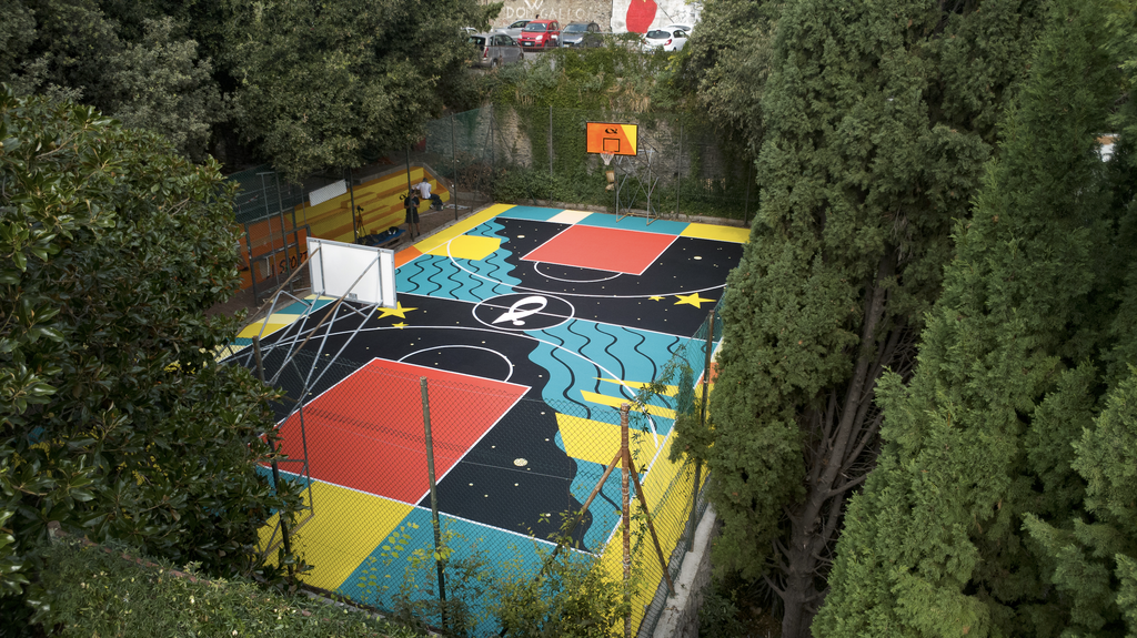 Vista dall’alto del playground SAB ai Giardini Buonvicini di Genova, immerso nel verde. Il campo da basket presenta una grafica geometrica e astratta con onde turchesi, stelle gialle, aree rosse e fondo nero.