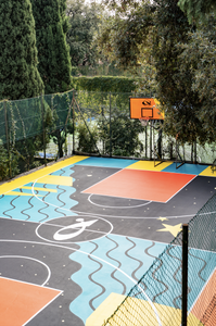 Dettaglio del canestro del playground SAB di Genova. In primo piano la pavimentazione rossa, gialla e nera; sullo sfondo le gradinate colorate con scritte dedicate allo sport e alla socialità.