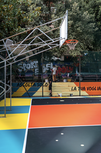 Vista laterale del campo da basket riqualificato da SAB a Genova, circondato da alberi. La pavimentazione alterna superfici nere e rosse, attraversate da onde turchesi, forme gialle e linee di gioco bianche.