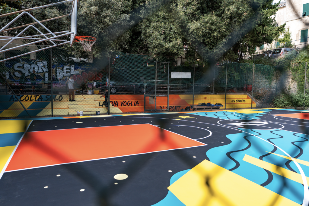 Vista laterale del playground SAB ai Giardini Buonvicini di Genova, con la grafica di Cabot Covet che combina onde turchesi, stelle gialle e aree rosse su fondo nero. Sullo sfondo, il canestro, le gradinate colorate con scritte dedicate allo sport e il logo Cisalfa, circondati dalla vegetazione.